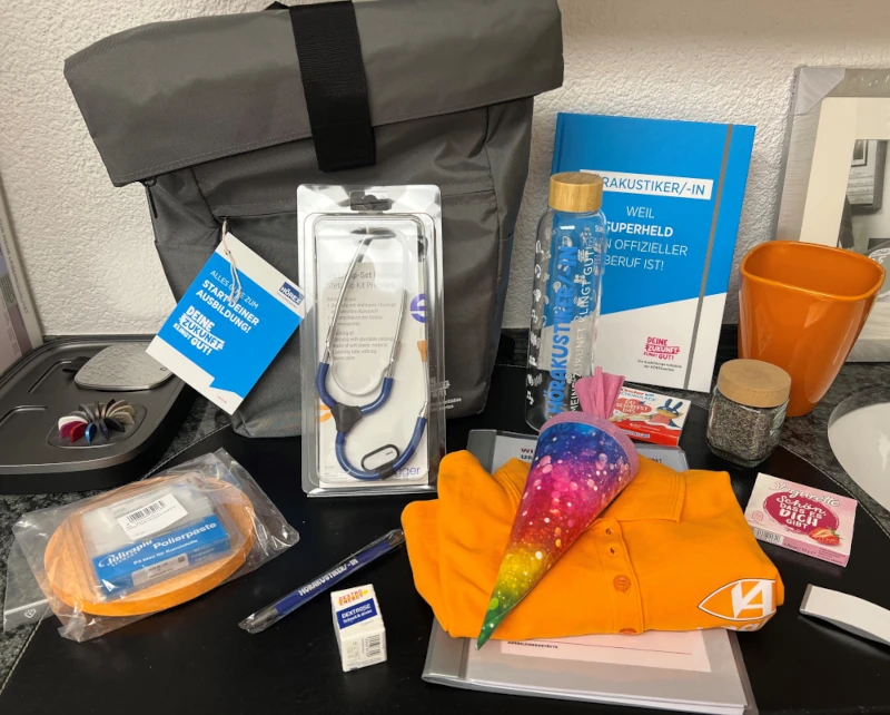 Verschiedene Geschenke für Hörakustiker-Ausbildung, darunter Rucksack, Wasserflasche und Schreibwaren.