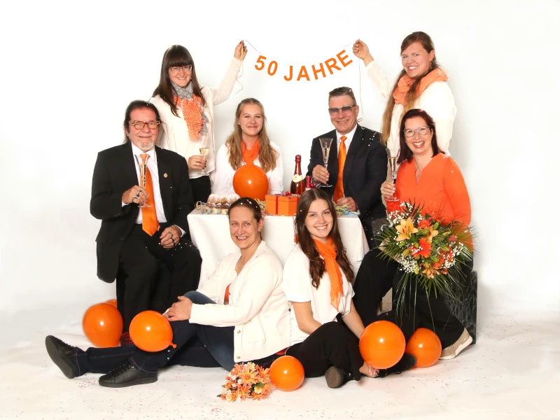 Gruppe feiert 50-jähriges Jubiläum mit Sekt, Blumen und orangefarbener Dekoration.